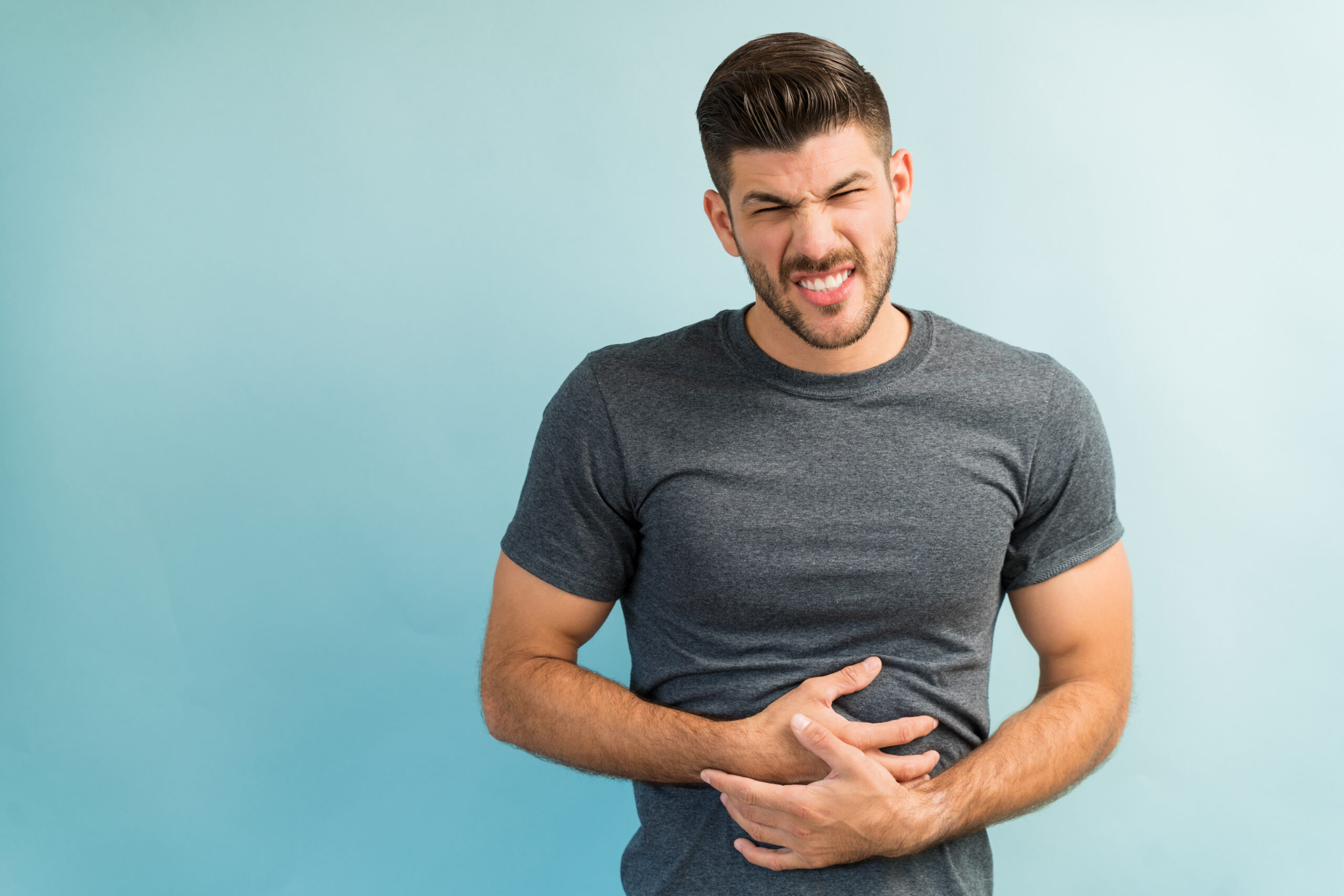 The image shows a young man clutching his stomach and frowning in discomfort, possibly indicating abdominal pain or digestive issues.