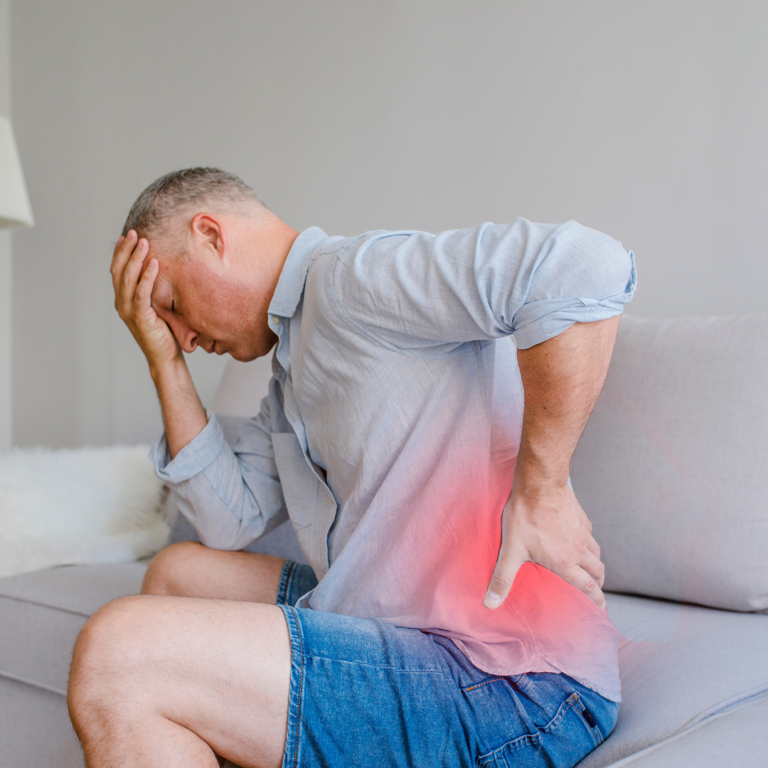 Man sitting on a sofa holding his lower back, experiencing back muscle pain and discomfort requiring natural pain relief.